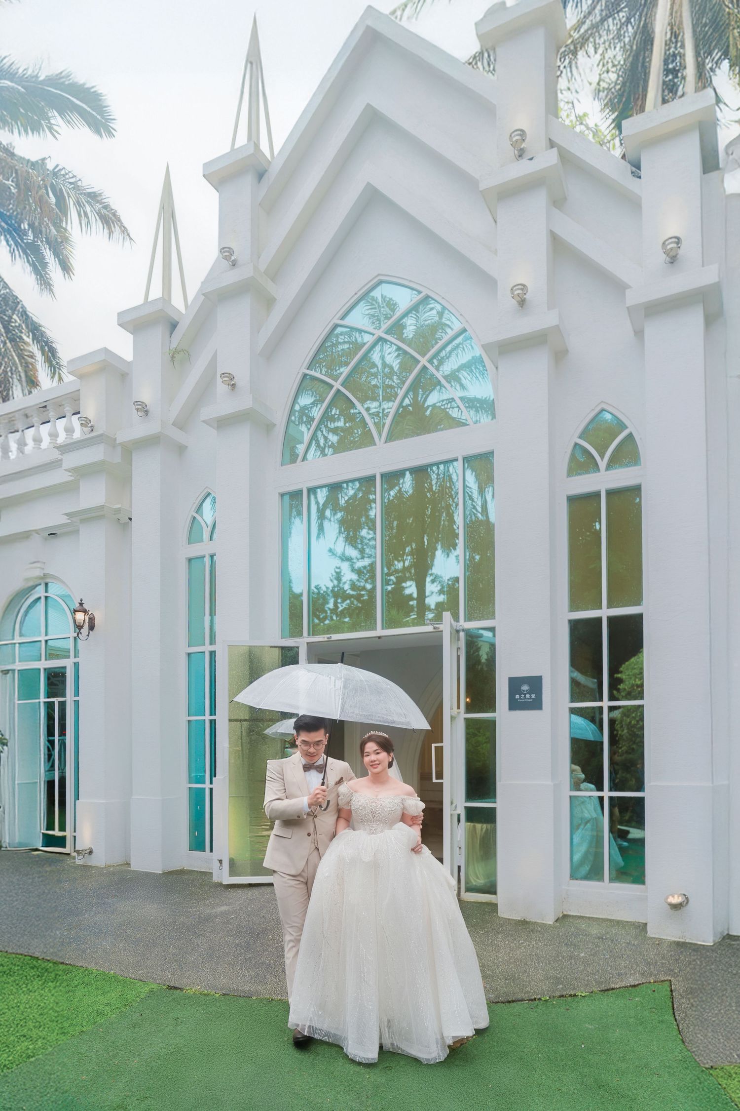 Elegant white chapel with arched turquoise windows and palm trees features a couple sharing an umbrella on a green carpet.
