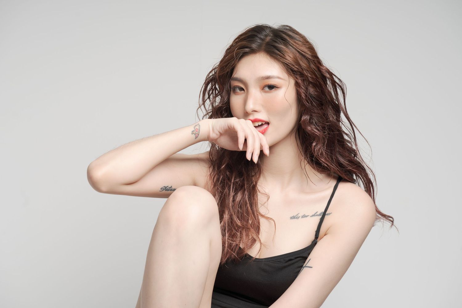Portrait of a woman with long wavy brown hair wearing black tank top against gray background, striking a casual pose.
