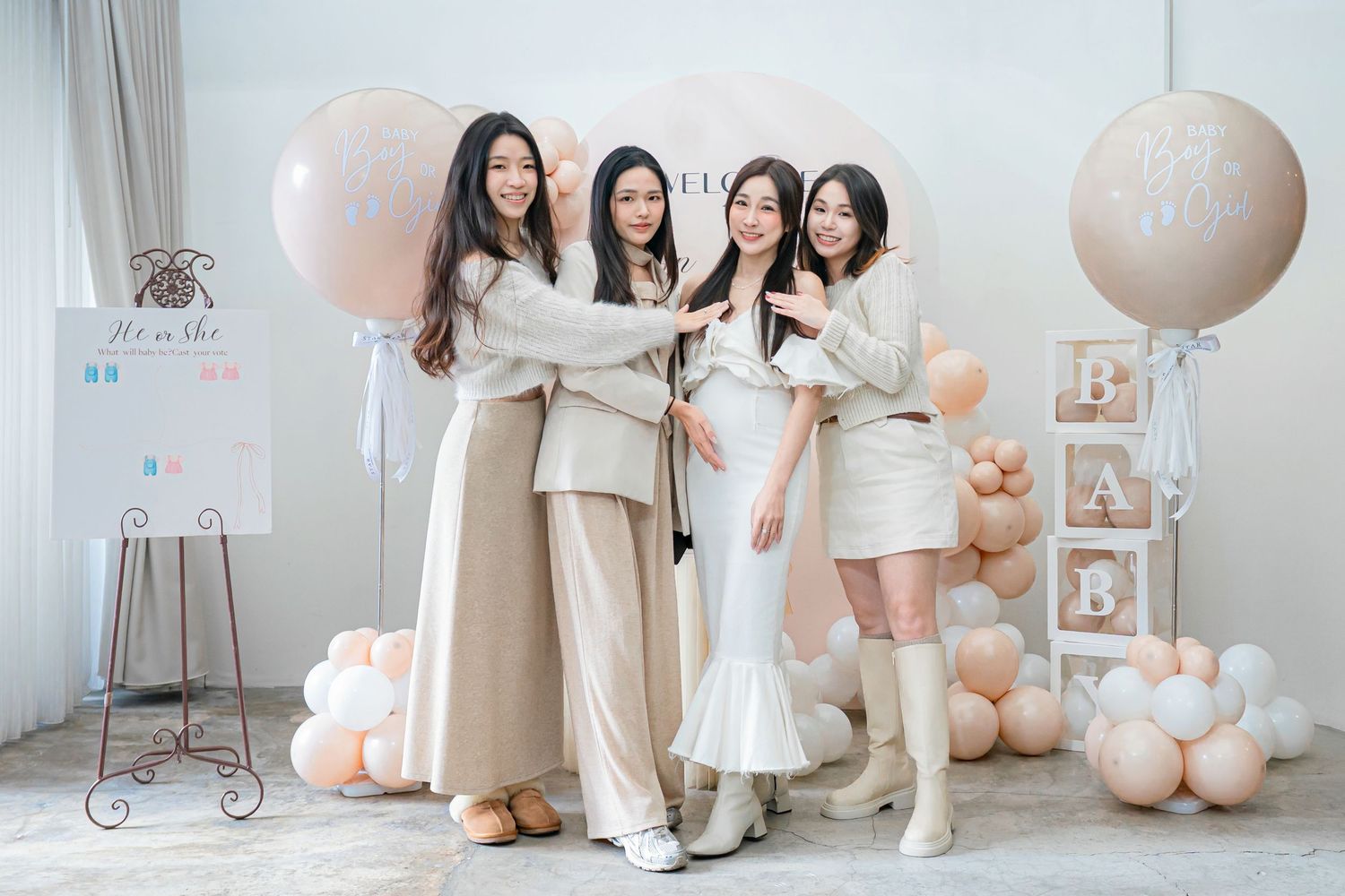 A group in neutral-toned outfits poses together at a pastel-decorated party setting with balloons and decorative elements.