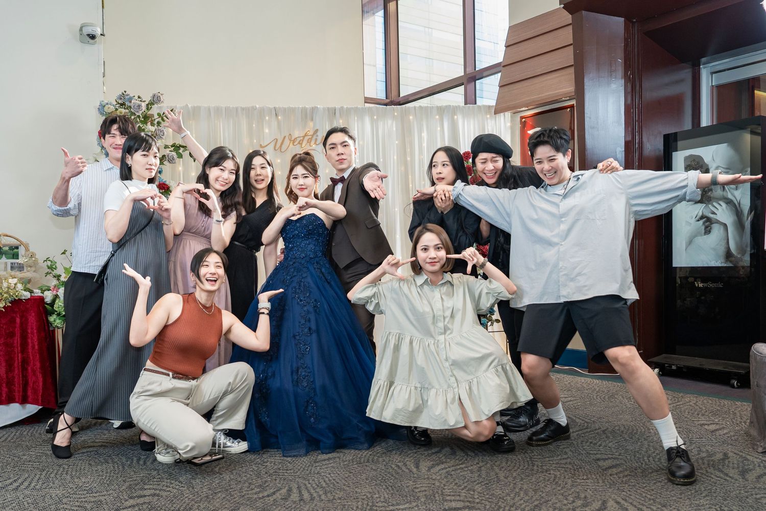 A group in formal and casual attire poses together at a celebration event, making playful gestures and expressions.