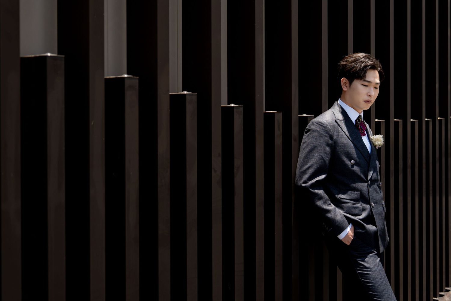 A well-dressed Asian man in a gray suit walks confidently past dark wooden architectural columns.