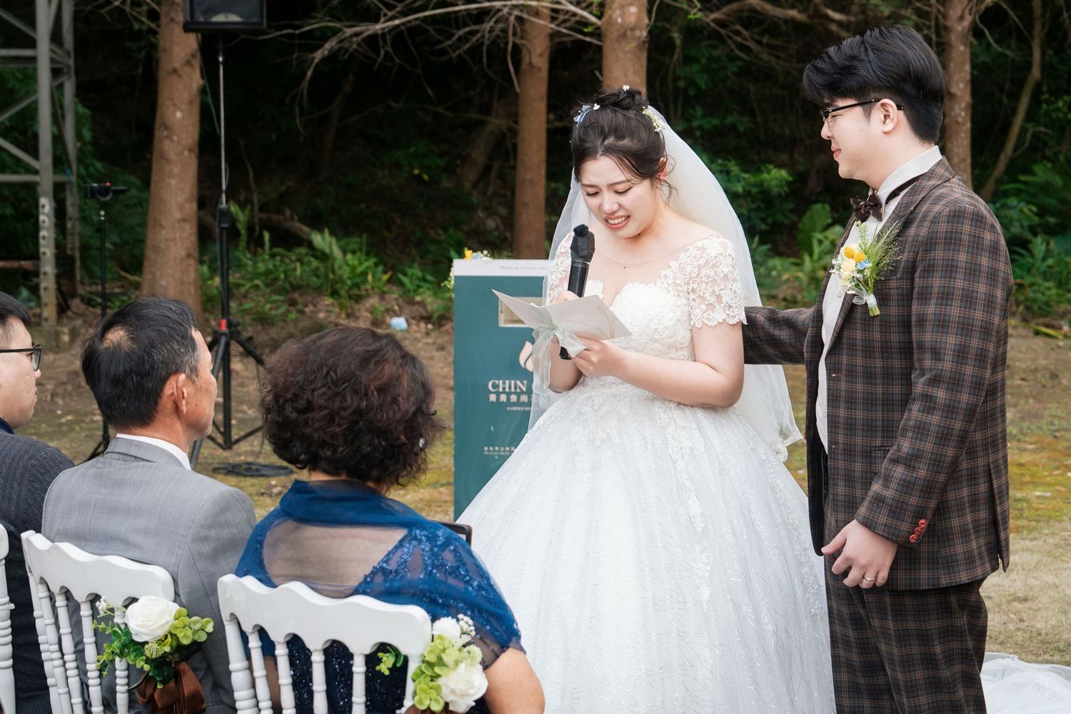 A bride and groom share a moment during an outdoor wedding ceremony with guests seated in white chairs.