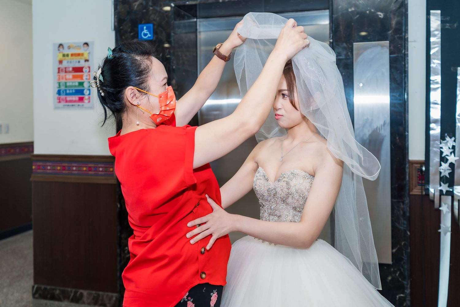 An assistant in red adjusts a bride's delicate white veil during wedding preparations.