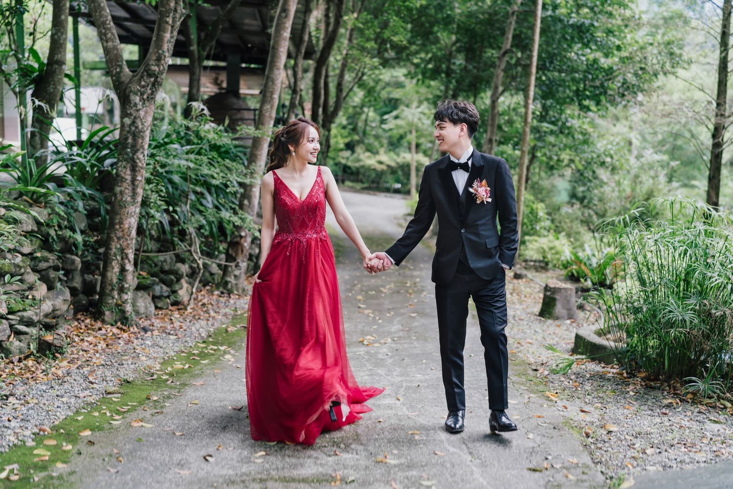 A couple walks hand in hand along a forest path, with the person in a flowing red gown and the other in formal black attire.