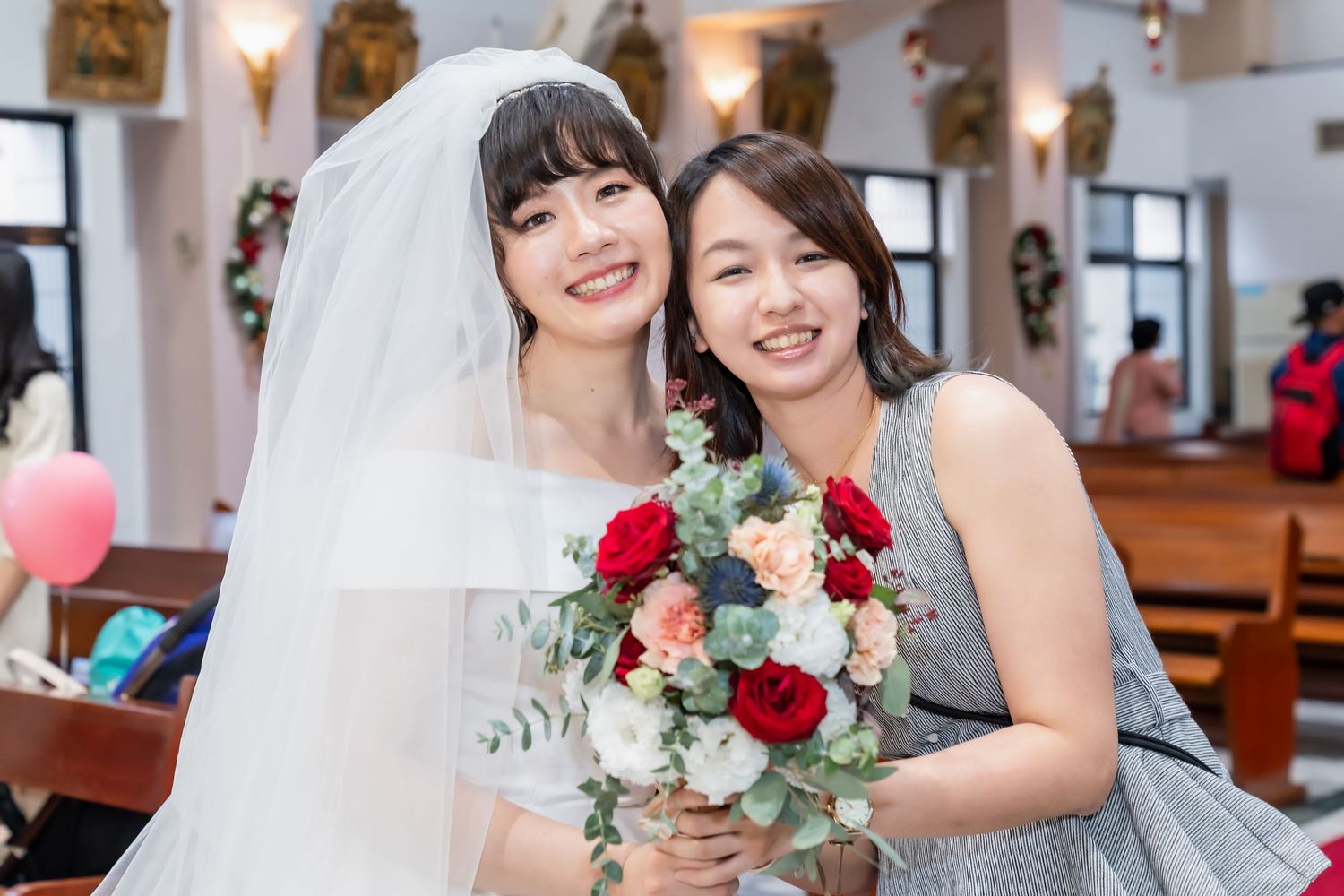 Two women embrace and smile while holding a bouquet of red and white flowers.
