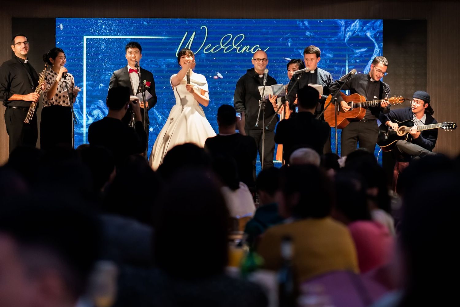 Wedding band performs on stage with instruments while guests watch from tables below.