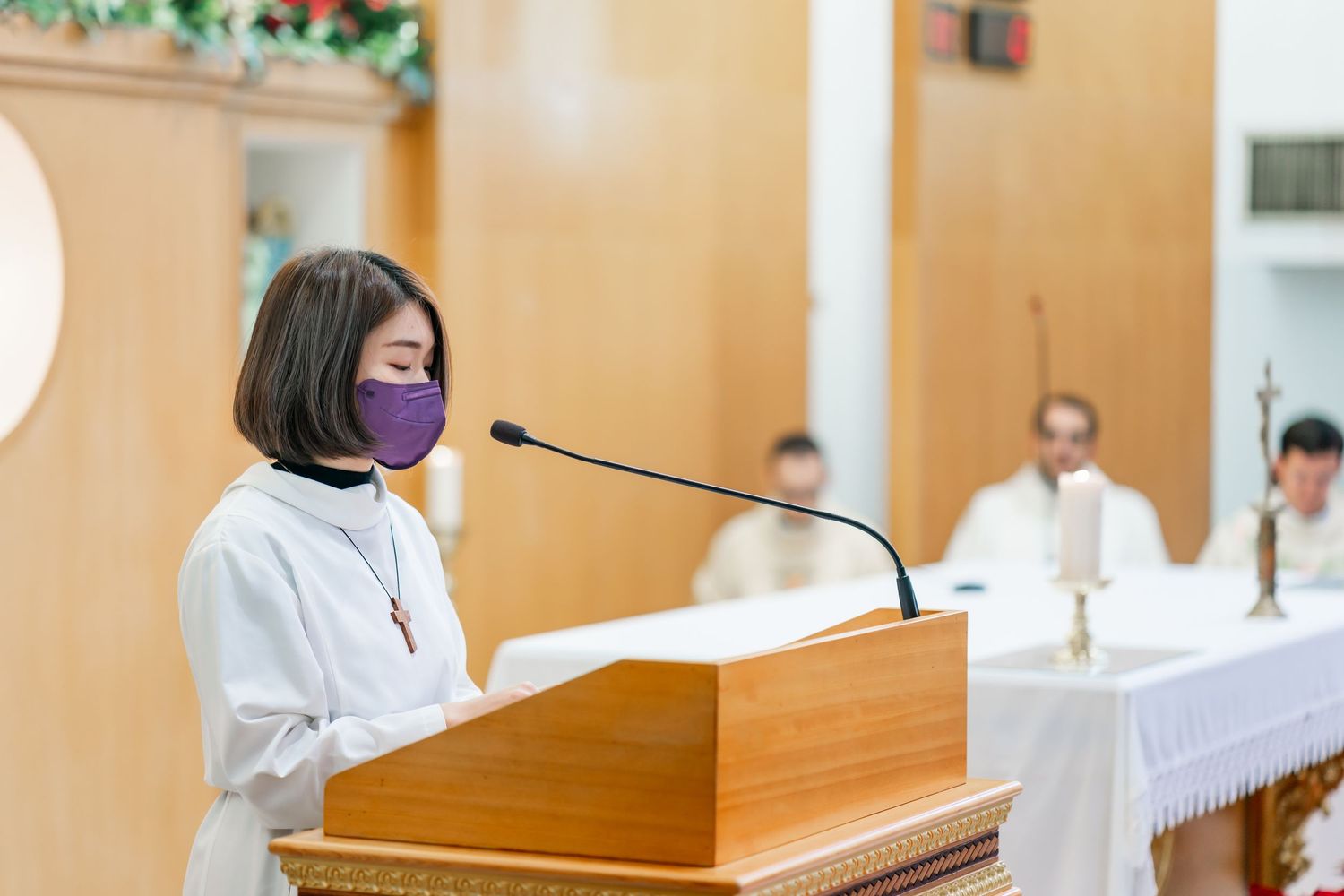 Speaker in white robe and purple mask addresses congregation from wooden podium.