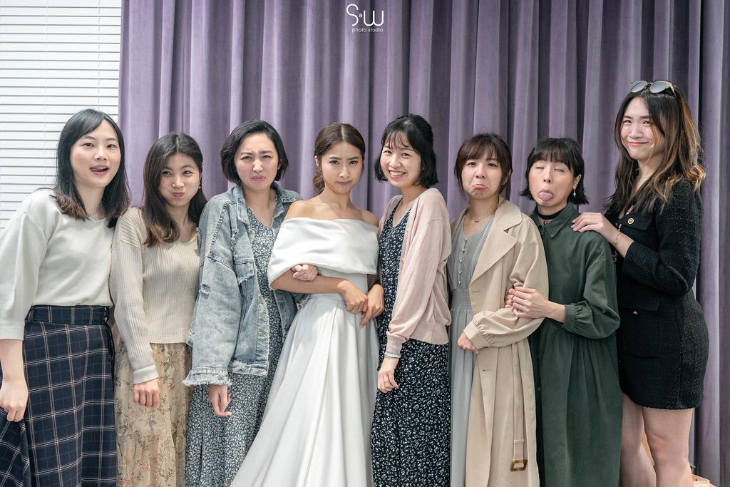 A bride in white off-shoulder gown poses with friends in casual attire against purple curtain.