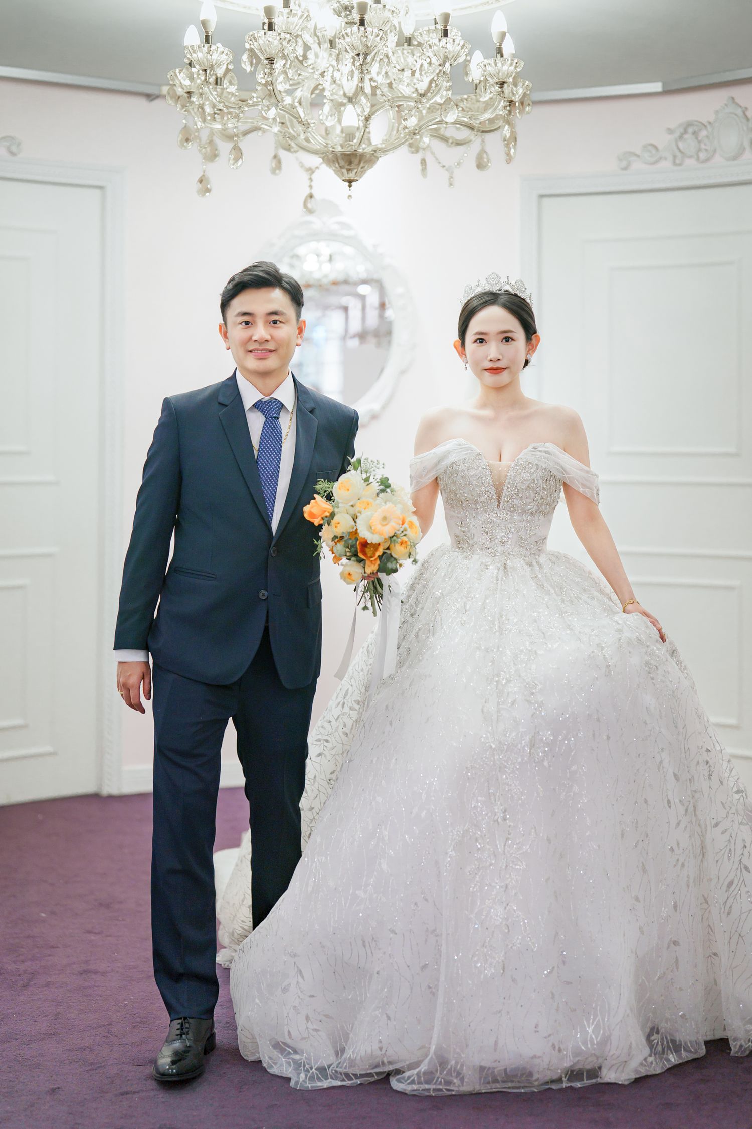 A couple in wedding attire poses together in an elegant white room with crystal chandeliers.