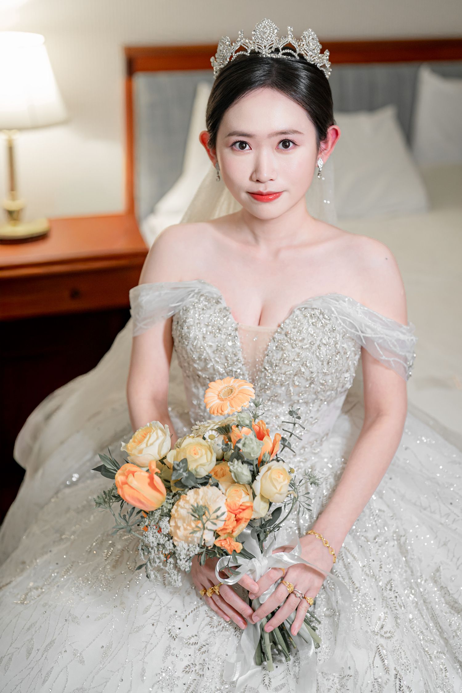 Bride in an ornate off-shoulder wedding gown with crystal tiara holds a peach and cream rose bouquet.