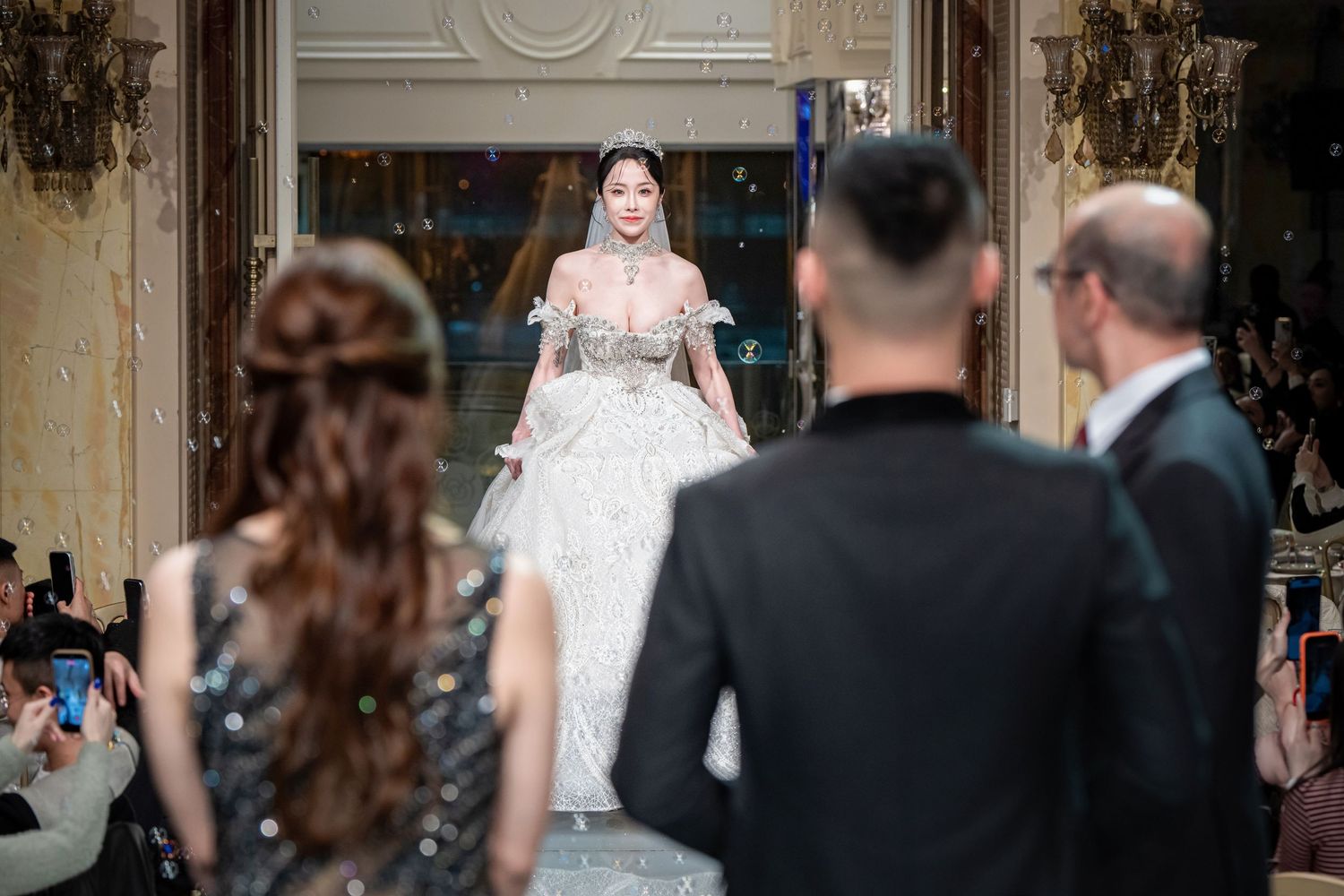 A bride in a white lace gown poses during a wedding ceremony in an ornate venue with elegant decor.