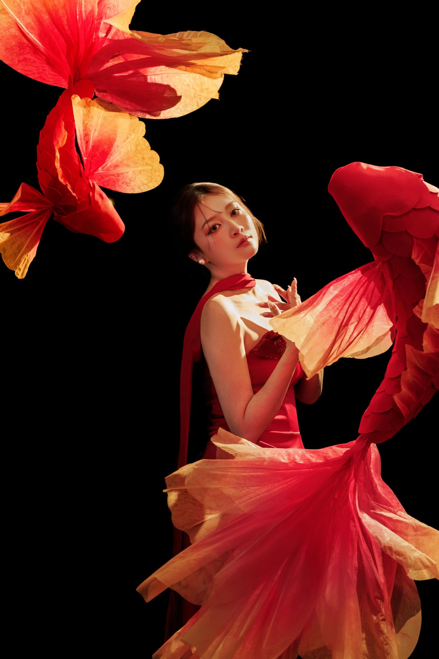 A graceful dancer in a flowing red and yellow dress poses elegantly against a black background with swirling fabric petals.