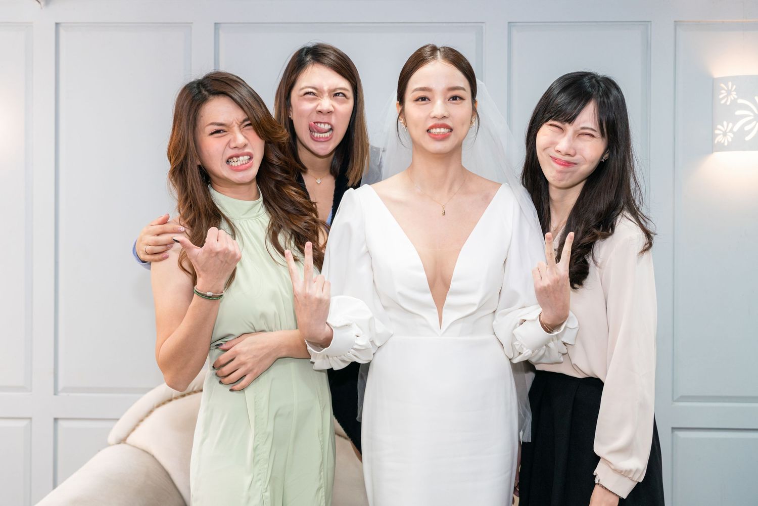 Four friends in pastel and neutral colored outfits pose together joyfully against a white wall at a special occasion.