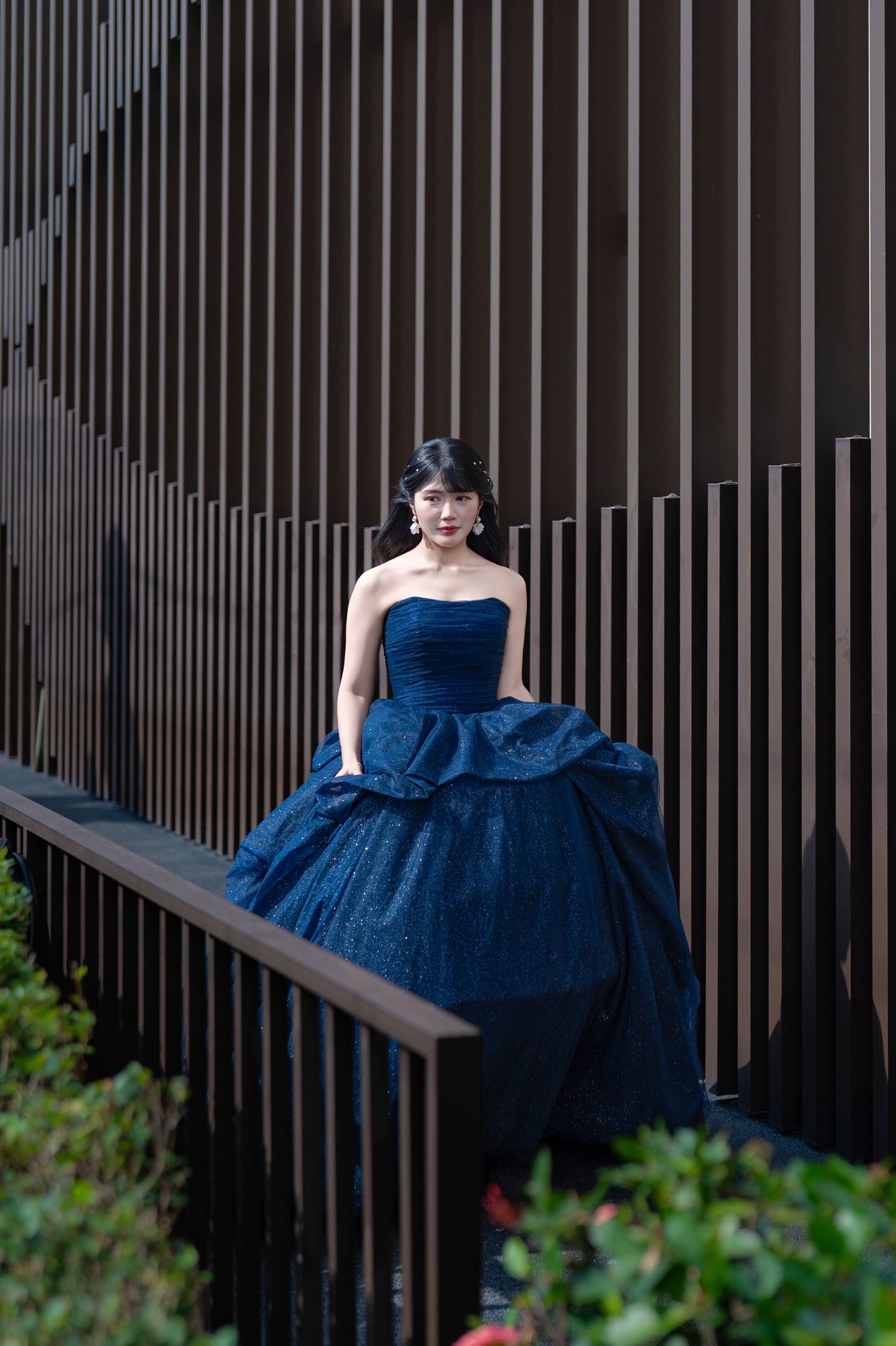 Woman in elegant navy blue strapless ball gown posing on stairs against modern dark wood slat wall.
