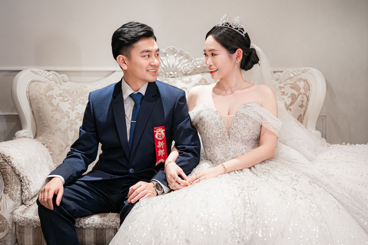 A couple in wedding attire sits together on an elegant white sofa, sharing a joyful moment during their celebration.