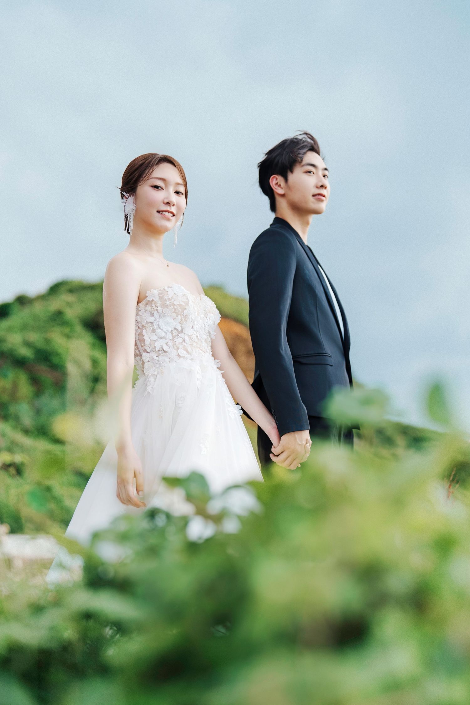 A bride in a white lace gown and groom in black suit pose together against a soft blue sky and green foliage backdrop.