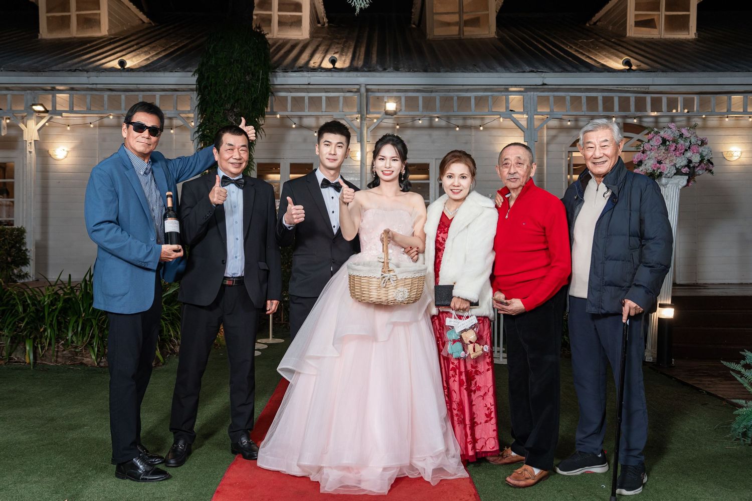 Wedding guests in formal attire gather outside a stone building for group photos at an evening celebration.