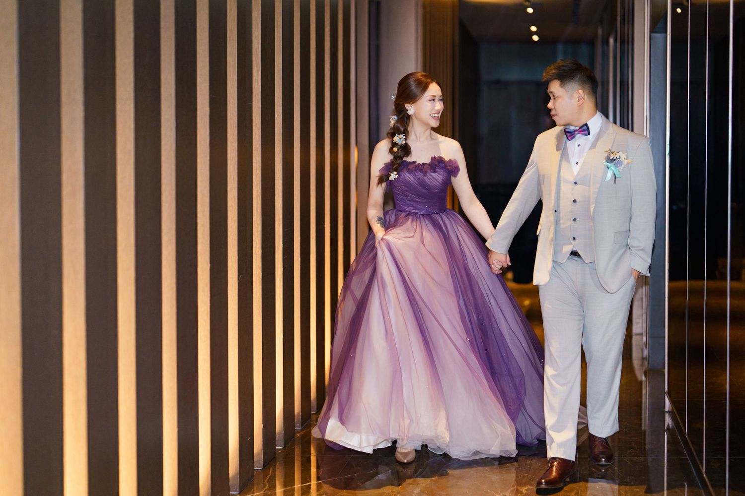 A couple in formal attire stands in front of gold vertical bars at an elegant event venue.