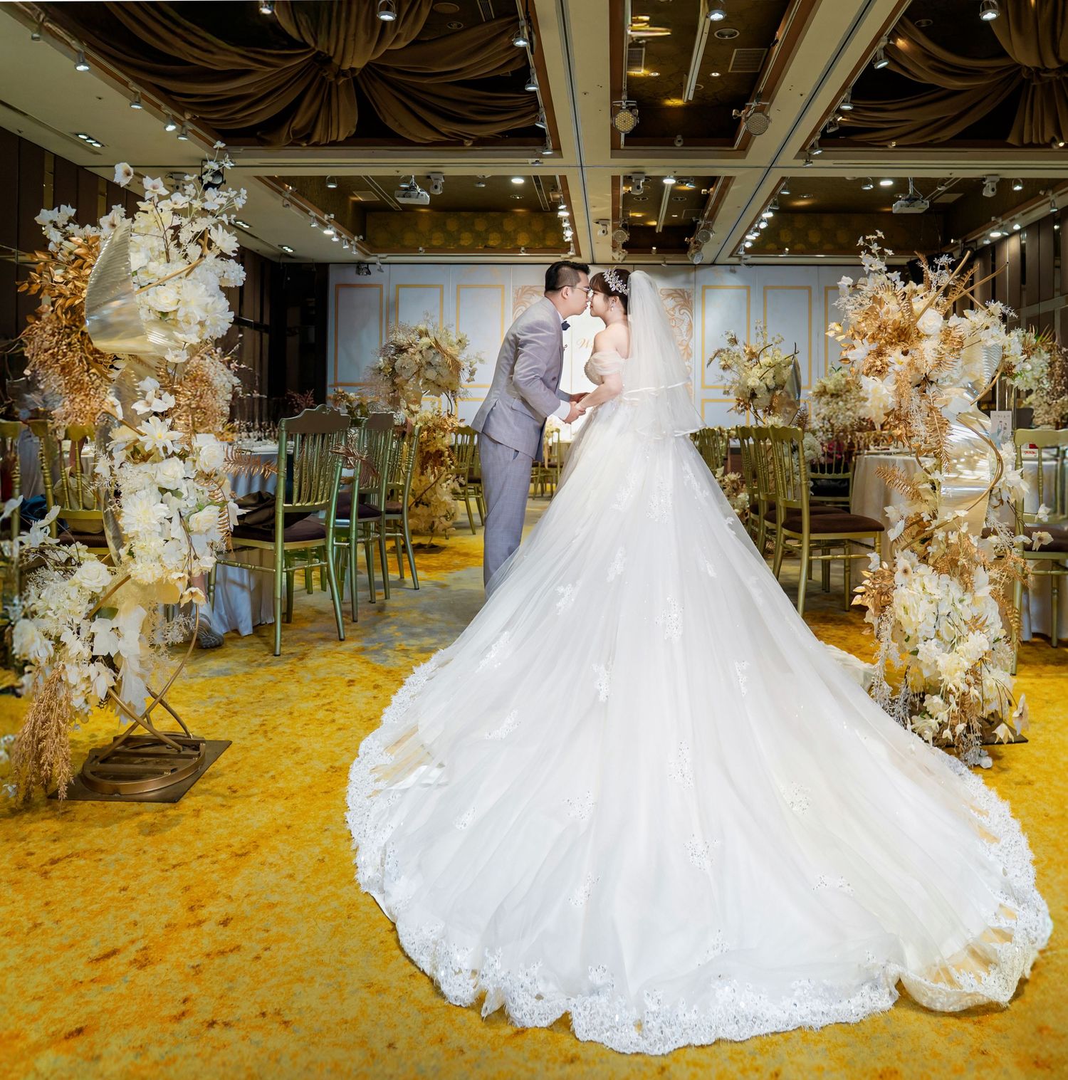 A couple in wedding attire dance together on a golden yellow carpet surrounded by elegant floral decorations.
