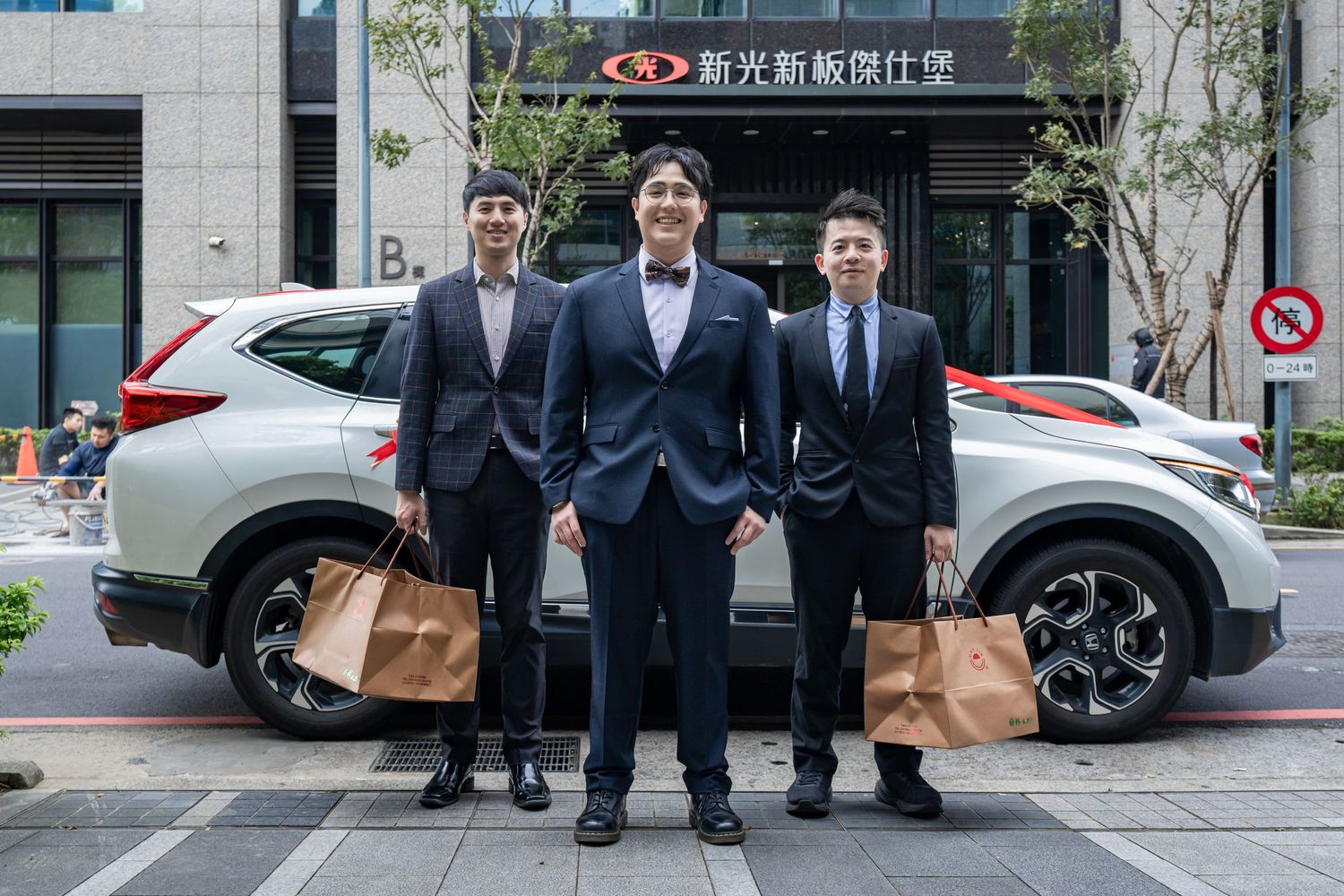 Four people in business suits walk together on a city sidewalk near parked cars and office buildings.