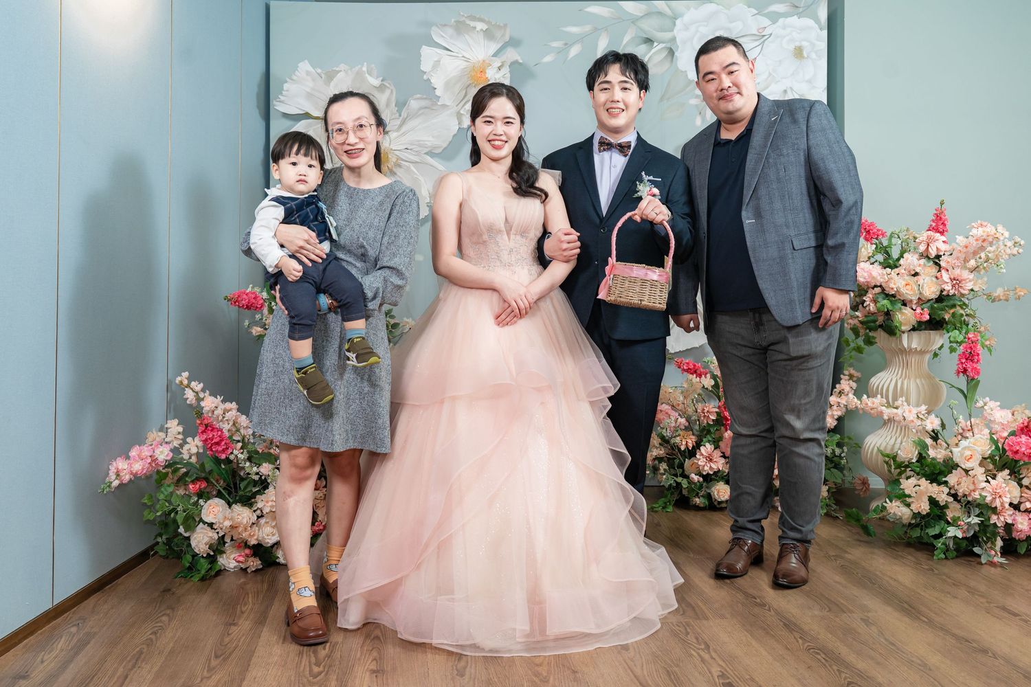 Wedding party poses for a series of formal portraits against a mint green wall with pink floral decorations.