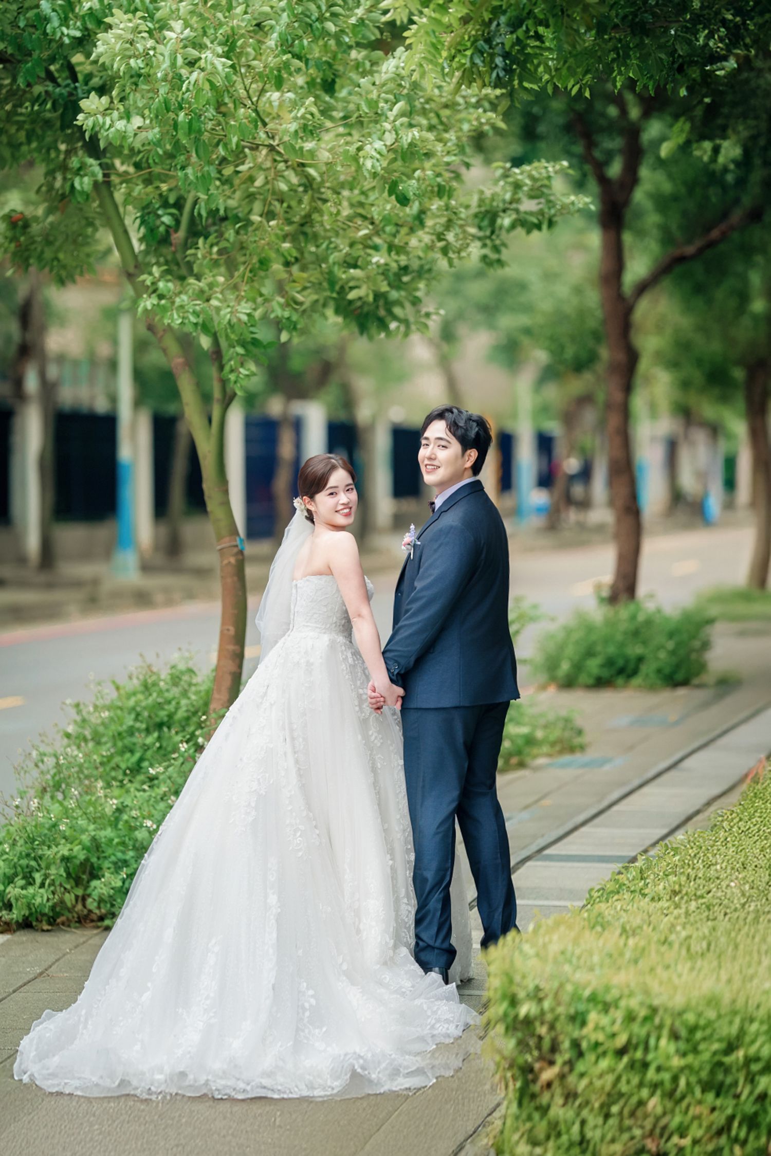 A couple in wedding attire holds hands while standing on a tree-lined pathway in an outdoor garden setting.