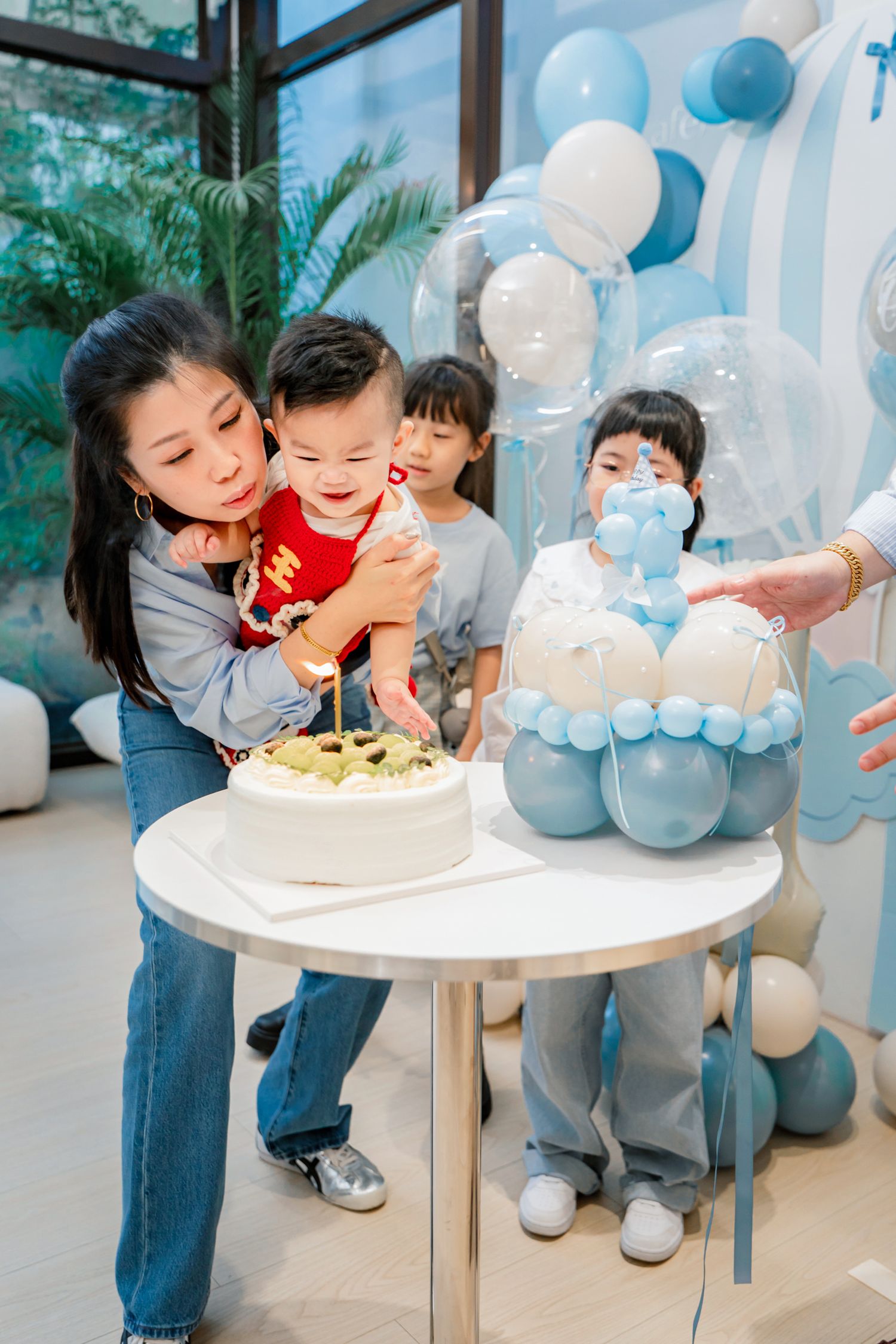 A birthday celebration with blue and white balloons as children gather around a white cake on a table.
