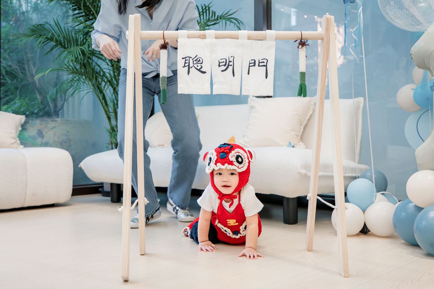 A small figure in a red outfit crawls under a wooden frame with Chinese calligraphy hanging above at a party setting.