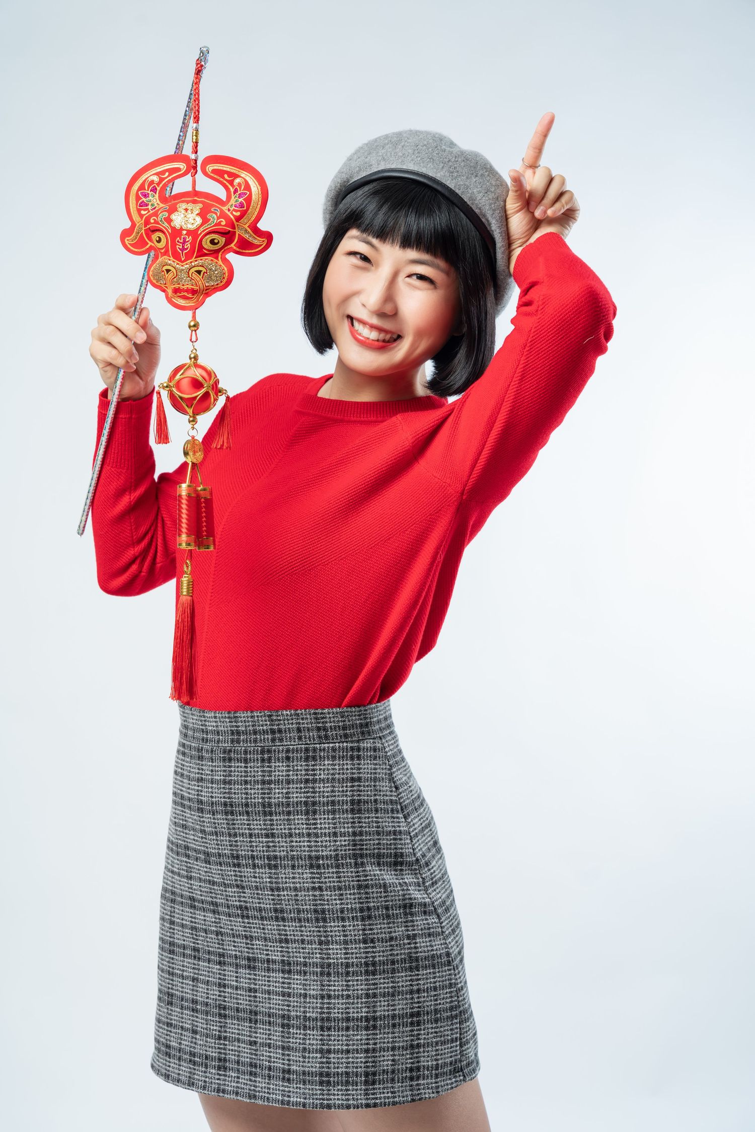 Person in red sweater and plaid skirt holds traditional Chinese New Year decorations in celebratory poses.