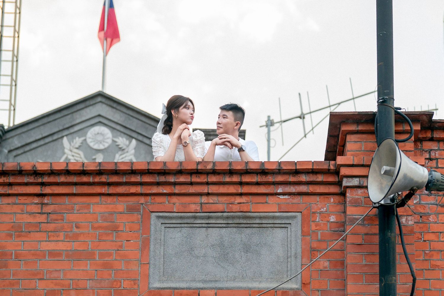 A couple shares a romantic moment on a brick rooftop with a red flag and satellite dish visible in the background.