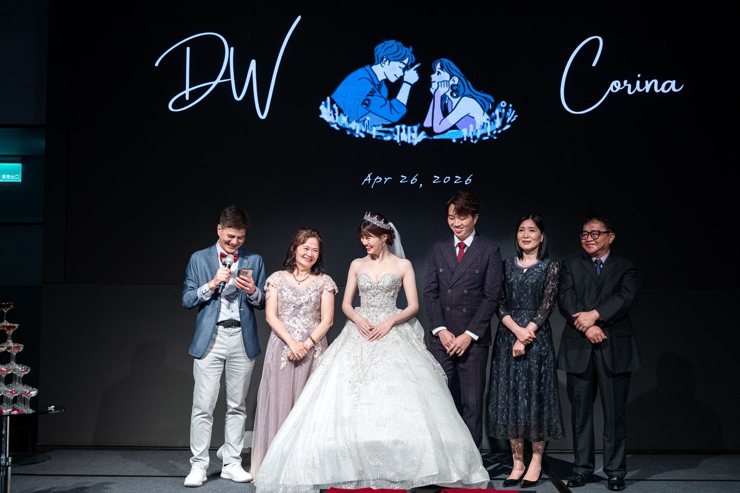 Wedding reception with bride in white gown and groom in suit posing with guests under DW and Corina April 2024 backdrop.