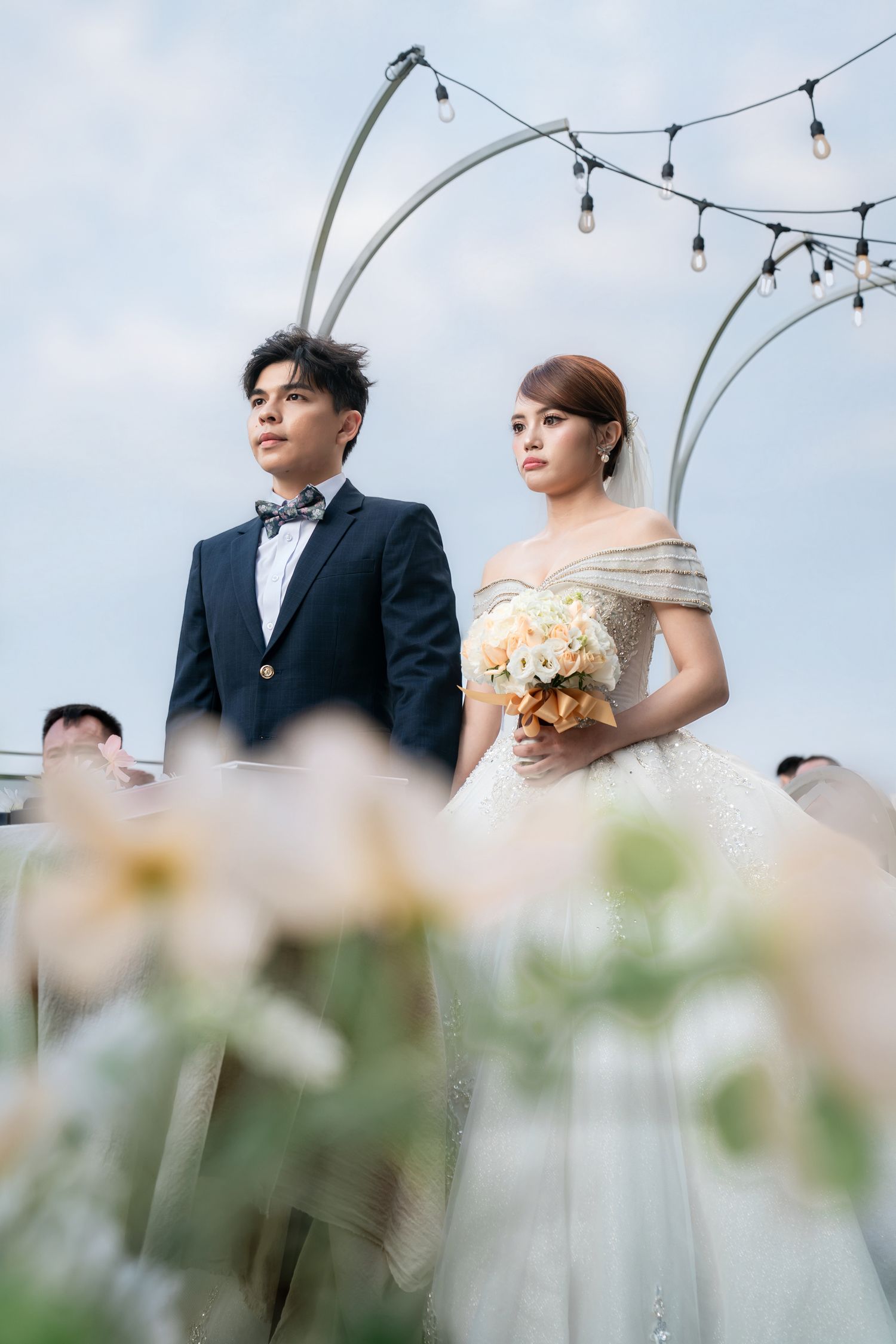 A bride in a white gown holding a bouquet stands beside a groom in a navy suit at an outdoor wedding ceremony.