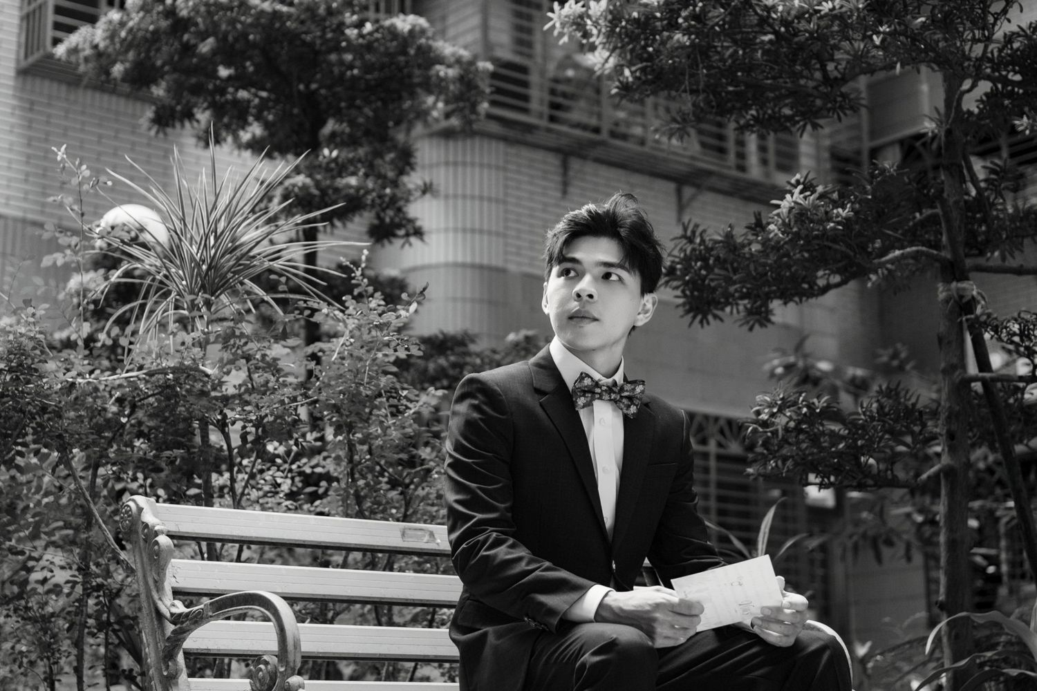 Young man in tuxedo with bow tie sits on bench holding papers, looking up thoughtfully in urban garden setting.