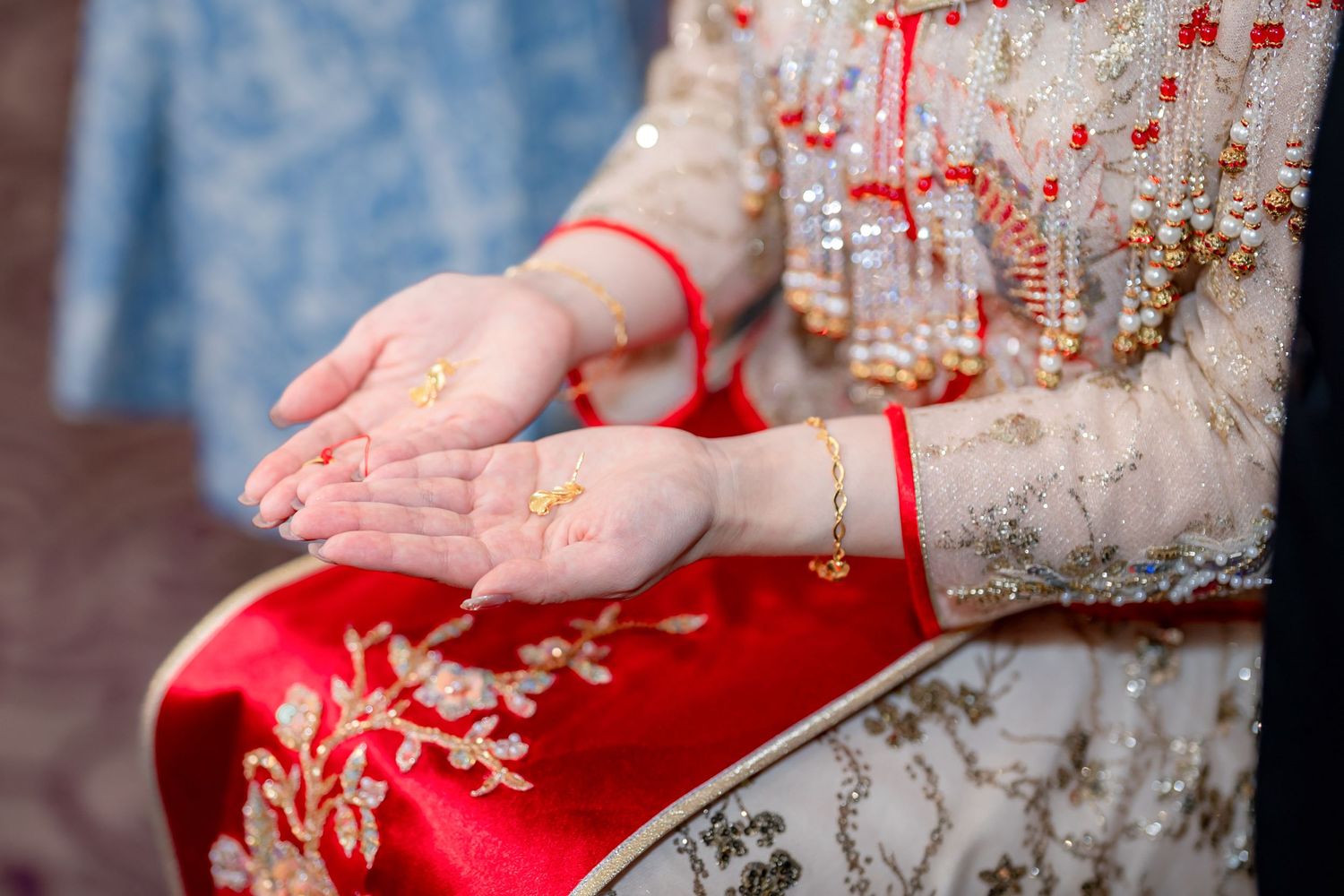 Bride's hands held palms up with henna and gold jewelry, wearing a red and gold embroidered outfit with beaded embellishments.
