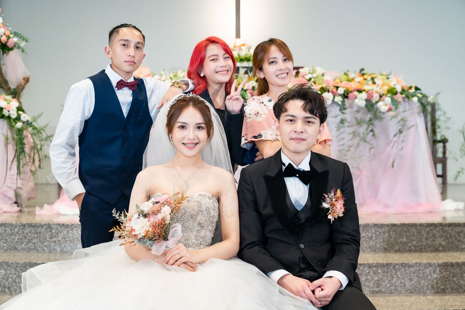 Wedding party poses together on church steps with floral decorations and pink backdrop.