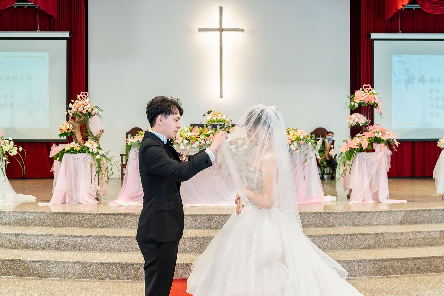 A wedding ceremony in a church with a illuminated cross and pink floral decorations.