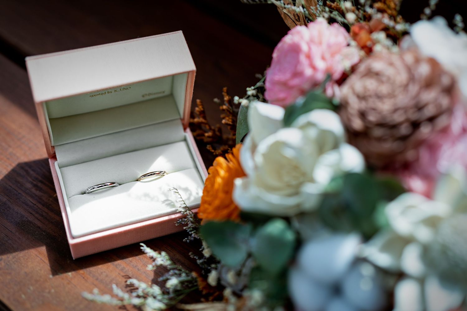Wedding ring box nestled among pink and white flowers on a rustic surface.