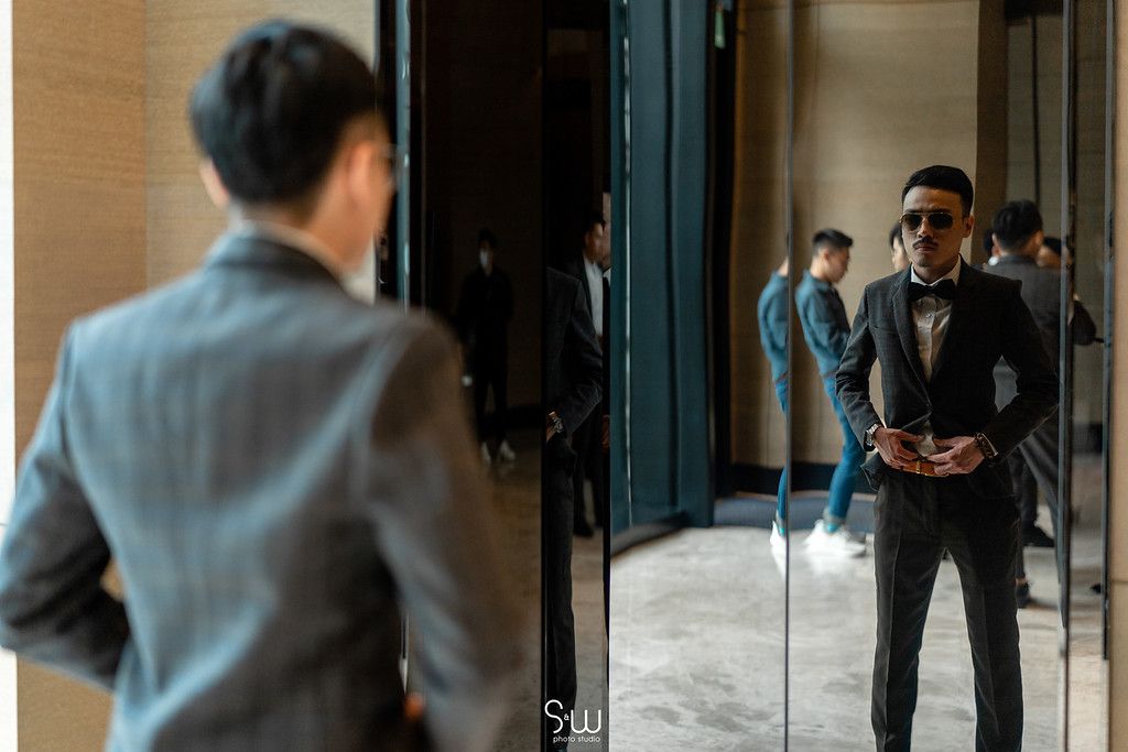 A man in a suit adjusts his bow tie while looking in a mirror.