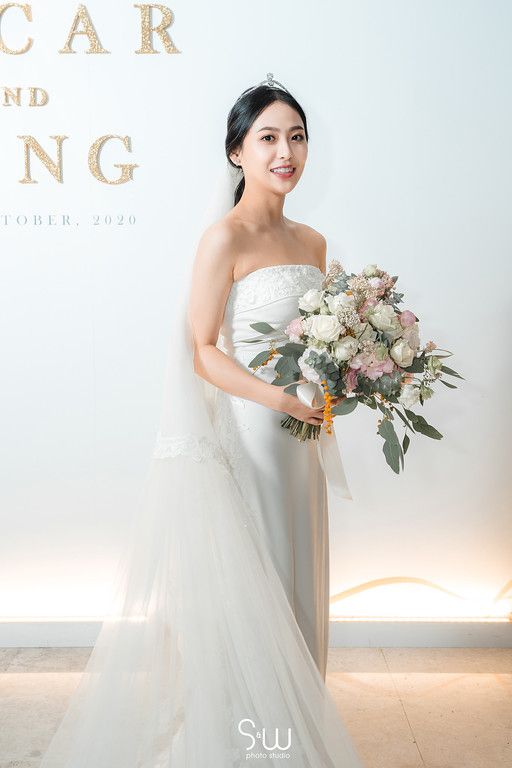 Bride in strapless white gown holds luxurious pastel flower bouquet against light background.