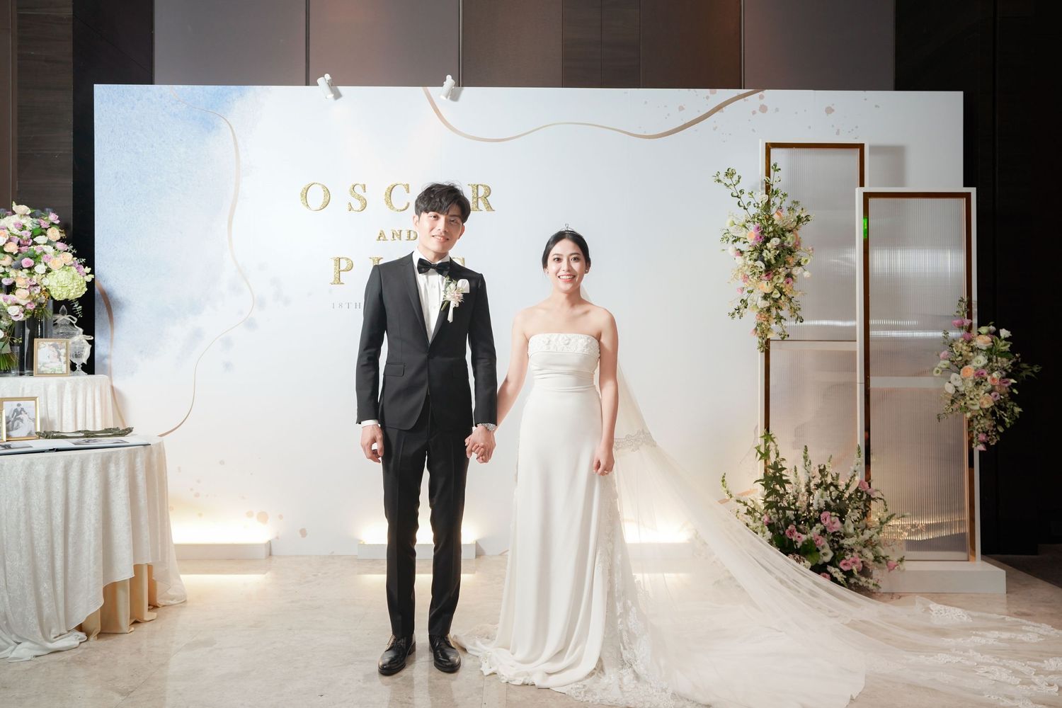 Elegant wedding couple stands before white backdrop with gold lettering and floral decorations.