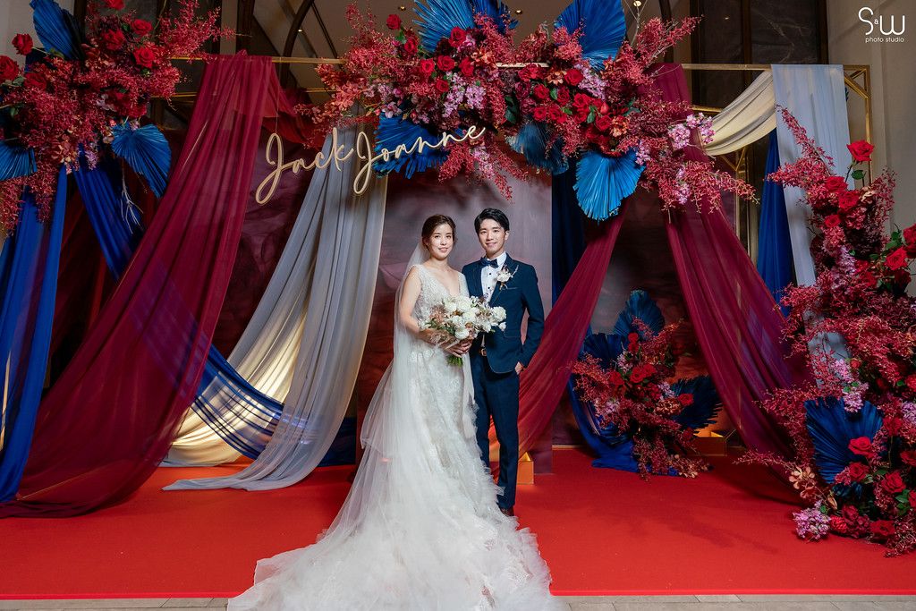 Wedding portrait under decorative arch with red flowers and blue draping.