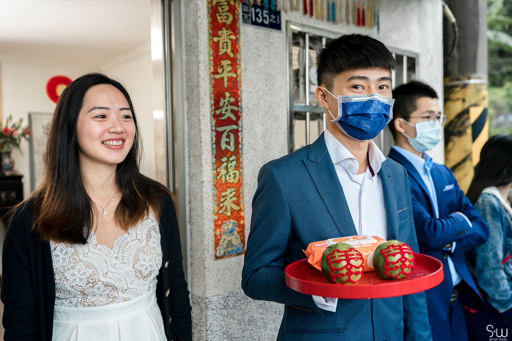 A couple stands in front of a building with traditional Chinese decorations as one holds red items on a tray.