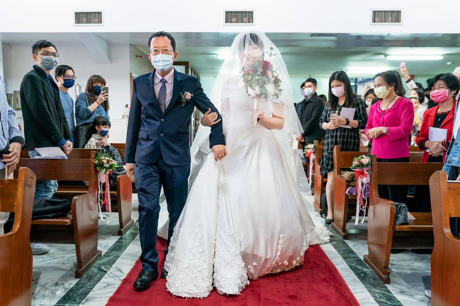 Wedding couple walking down red carpet aisle in church while guests observe from pews.