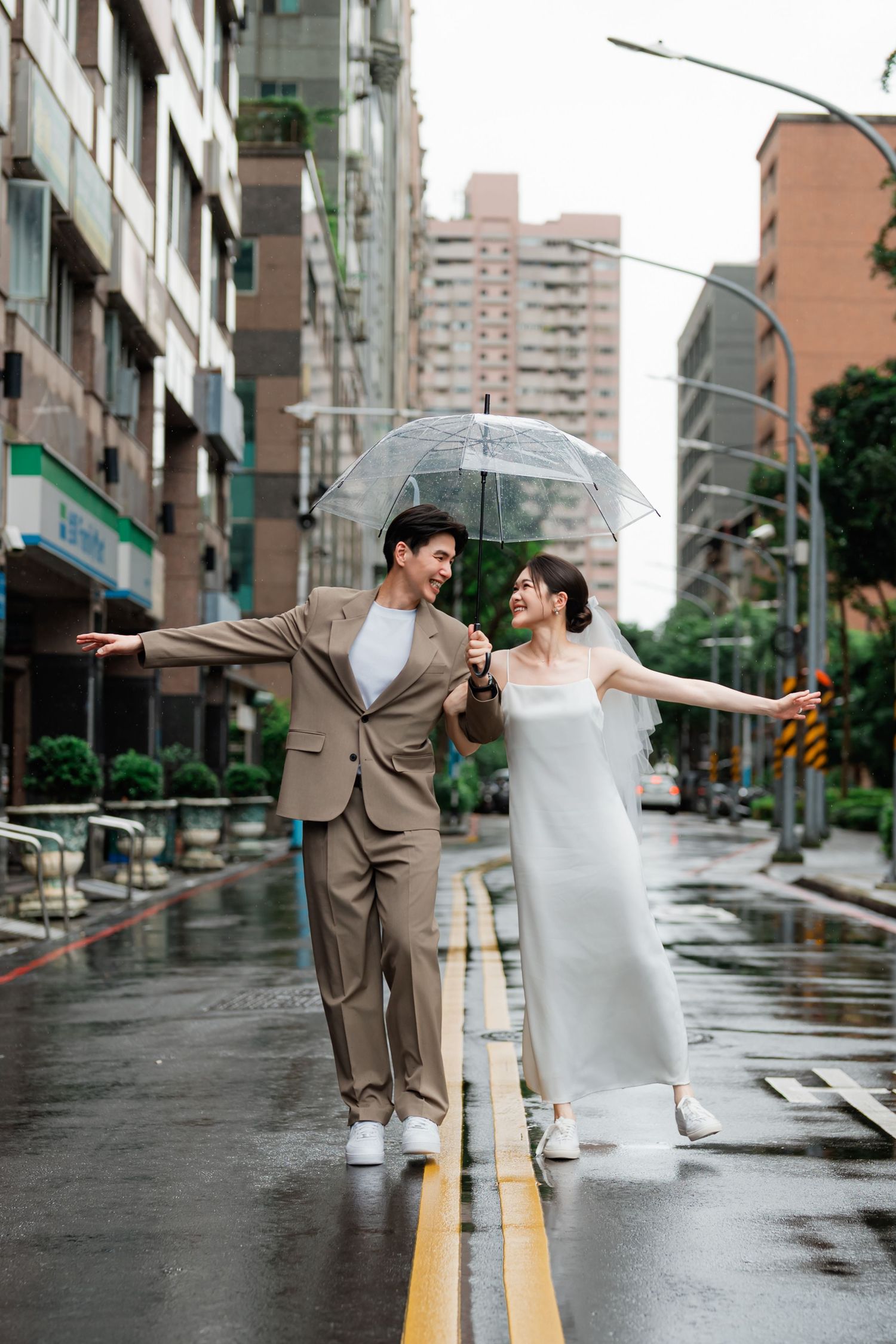A couple balancing on a wet street divider while sharing a clear umbrella on a rainy day in the city.