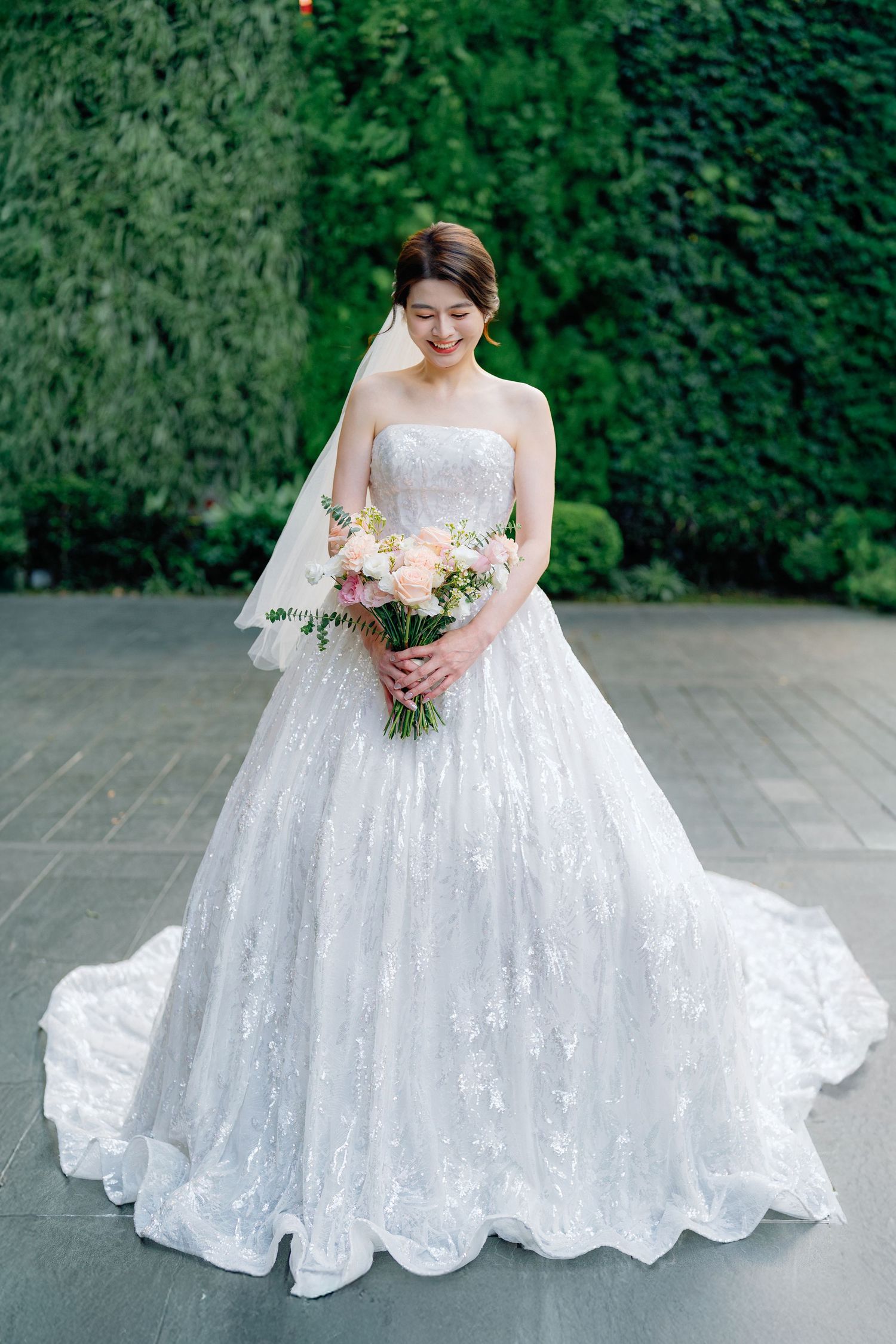Stunning white lace wedding gown with long train and strapless neckline captured against lush green garden backdrop.