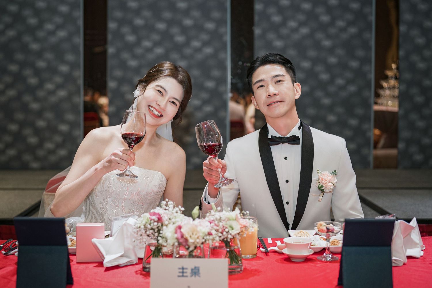 Wedding reception table with people holding wine glasses and toasting over elegant red and white decor.