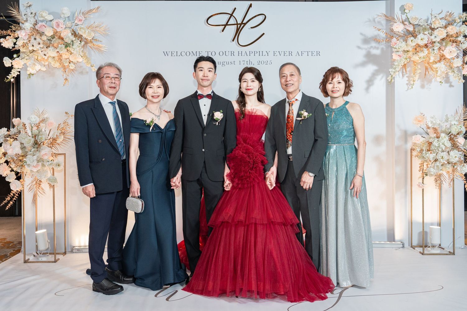 Formal group photo at elegant wedding celebration with decorative floral arch and bride in stunning red gown.