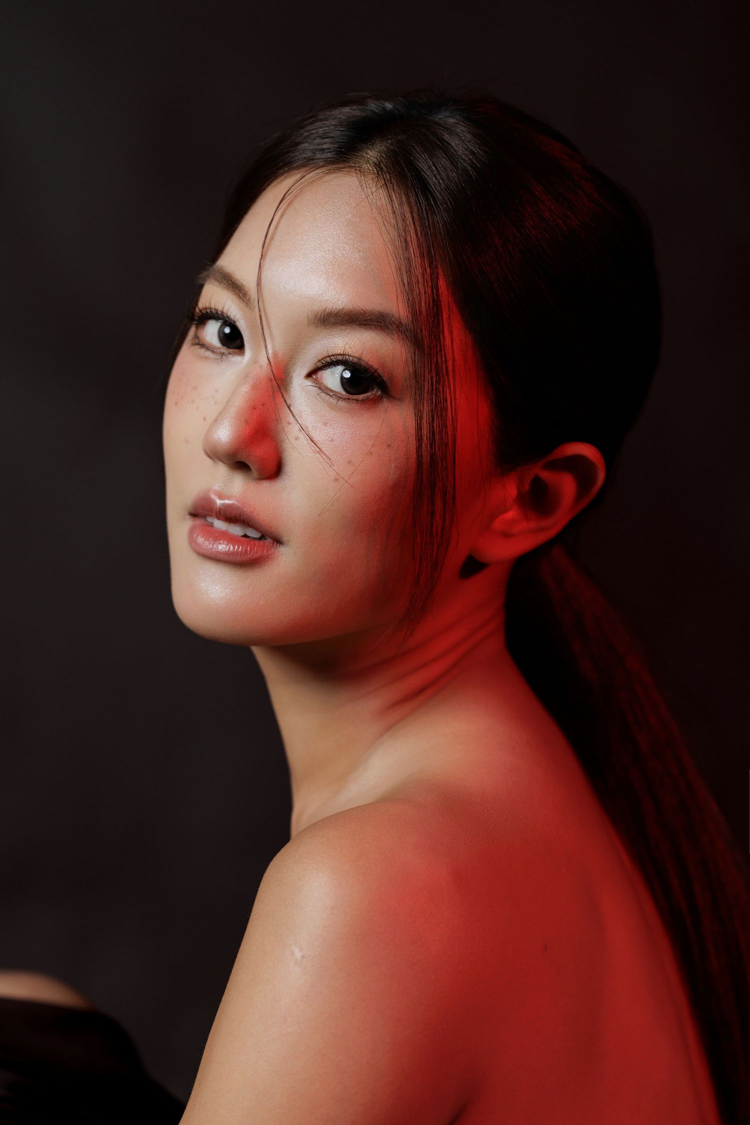 Portrait of a young Asian woman with red dramatic lighting, sleek ponytail, and natural makeup on dark background.