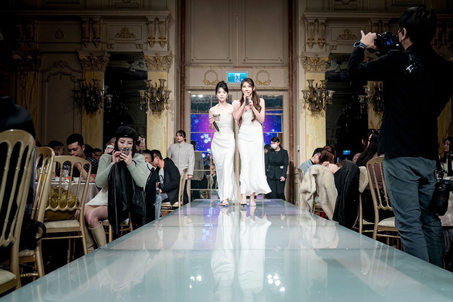 Models walk the runway in elegant white gowns during a bridal fashion show in an ornate venue.