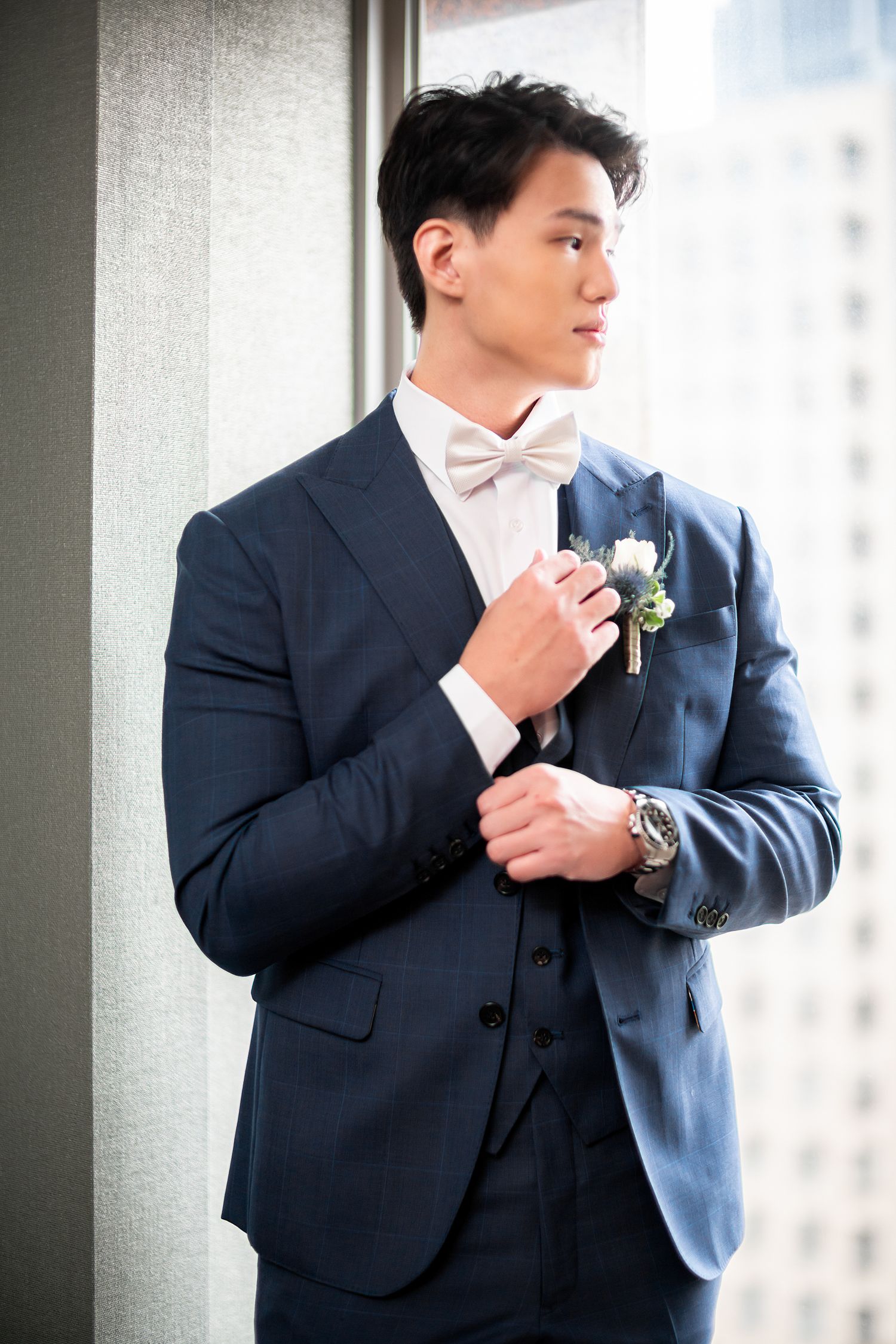 Elegant groom in navy blue suit and white bow tie adjusts boutonniere while standing by window.
